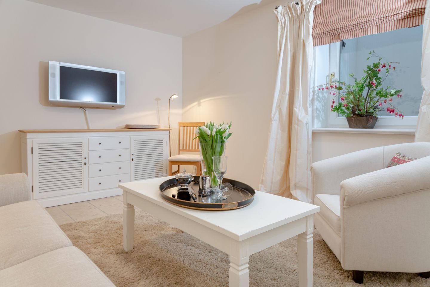 Ein Wohnzimmer mit einem an der Wand montierten Flachbildfernseher, einem weißen Couchtisch mit Tablett, einem weißen Sofa, einem weißen Sessel und einem Fenster mit einem Blumentopf.