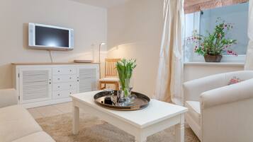 Ein Wohnzimmer mit einem an der Wand montierten Flachbildfernseher, einem weißen Couchtisch mit Tablett, einem weißen Sofa, einem weißen Sessel und einem Fenster mit einem Blumentopf.