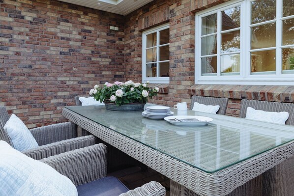 A glass-top table with wicker chairs and a bench, set against a brick wall with large windows.