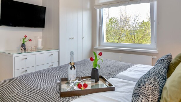 Une chambre à coucher avec un lit, une table de chevet sur laquelle se trouvent une bouteille et des fleurs, un téléviseur fixé au mur et une fenêtre donnant sur des arbres.