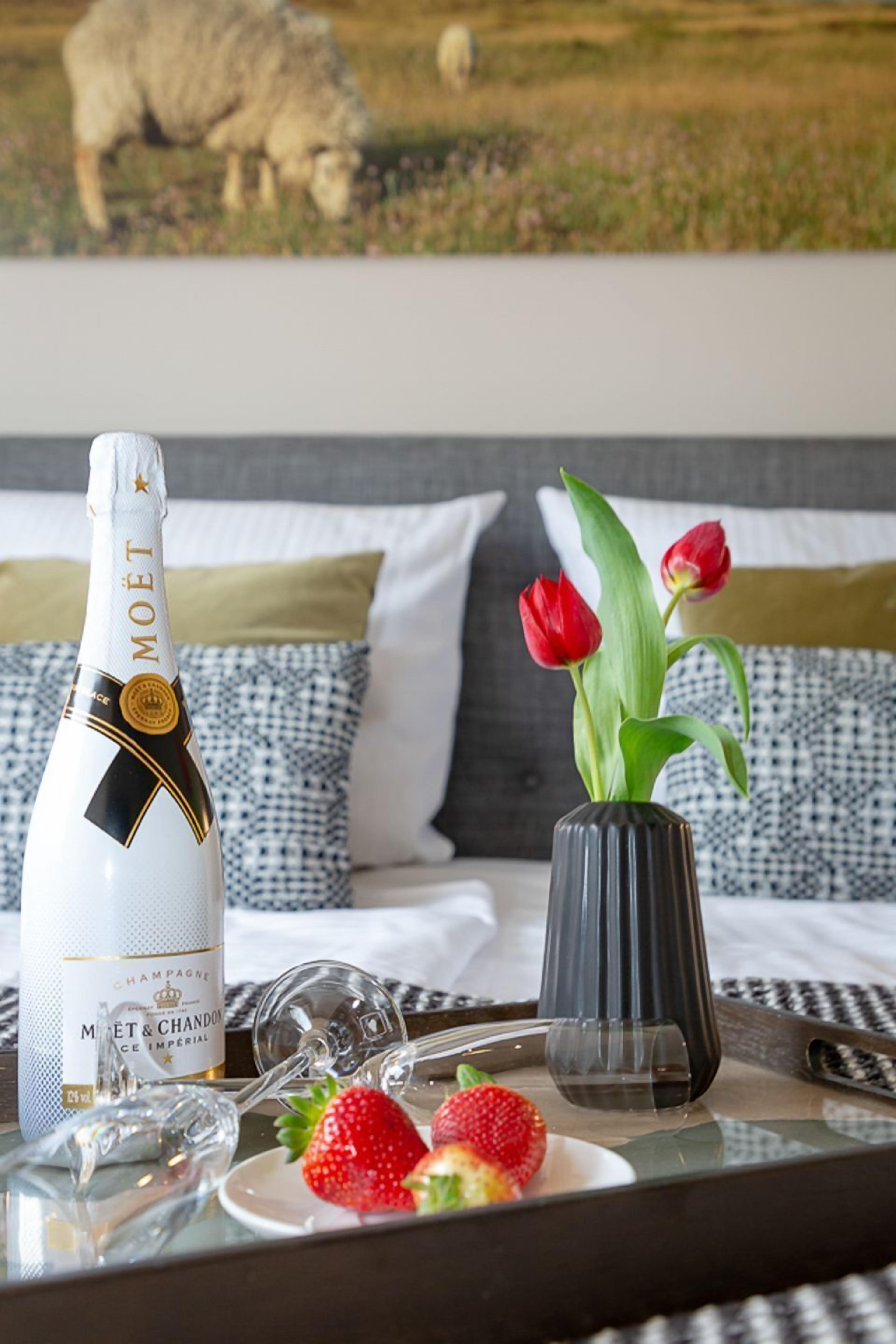 A bed with a white and black patterned pillow, a bottle of Moët champagne, a vase with red tulips, and a tray with strawberries and wine glasses.