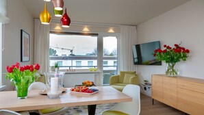 Un salon moderne avec un coin repas, comprenant une table en bois, des chaises blanches, un canapé, un téléviseur et un vase rempli de fleurs rouges.