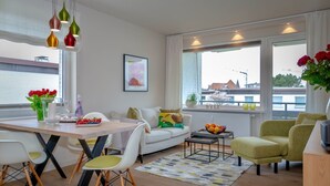 Un salon moderne comprenant un coin repas, des meubles blancs et de grandes fenêtres offrant une vue sur la ville.