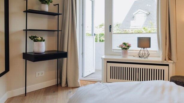 A bedroom with a bed, a window with curtains, a radiator, and two shelves with plants. - Poseidon - Poseidon: A luxurious retreat for 4 sea lovers (Sylt)
