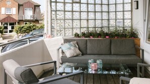 A balcony with a glass table, grey sofa, and chairs. There are potted plants on the windowsill and a view of a house in the background. - Poseidon - Poseidon: A luxurious retreat for 4 sea lovers (Sylt)