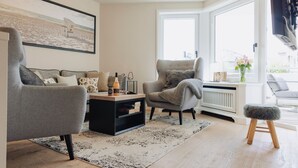 A modern living room with a grey sofa, a black coffee table, and a grey armchair. There is a window with a view of the outside, a radiator, and a small stool. - Poseidon - Poseidon: A luxurious retreat for 4 sea lovers (Sylt)