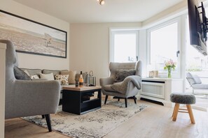 Una sala de estar moderna con un sofá gris, una mesa de centro negra y un sillón gris. Se ve una ventana con vistas al exterior, un radiador y un taburete pequeño.