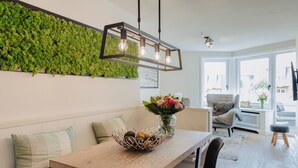 A modern living room with a wooden dining table, a bench with cushions, a wall-mounted green plant, and a hanging light fixture. - Poseidon - Poseidon: A luxurious retreat for 4 sea lovers (Sylt)