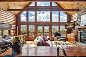A log cabin with a large open living area featuring a stone fireplace, wooden furniture, and a scenic mountain view through large windows. - VIEWS! Spacious and relaxing cabin! VIEWS! (Townsend)