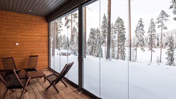 Terraza de madera con tres sillas y una mesa, con vistas a un bosque nevado a través de amplios ventanales.