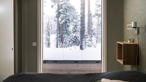 Un dormitorio con un ventanal grande, una mesita de noche con lámpara y vistas a árboles cubiertos de nieve.