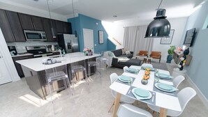 Une cuisine moderne dotée d’un îlot central, un coin repas avec une table blanche dressée pour quatre personnes, et un espace salon avec un canapé et une table basse.
