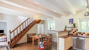 A spacious kitchen with wooden flooring, a dining area with a wooden table and chairs, a staircase with a wooden railing, and a hanging light fixture. - Ti ar Stang by Interhome (Esquibien)