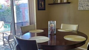 A round wooden dining table with four chairs, each with a woven placemat. A glass vase with a candle is on the table. - Sunland Cottage in 55+ Golf Community (Mesa)