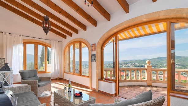A sunlit patio with wicker furniture, large arched windows, and a view of the countryside.