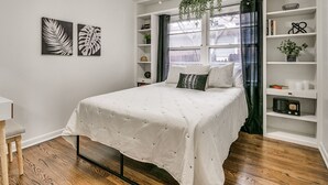 Un dormitorio con una cama, un ventilador de techo, una ventana con cortinas y una estantería.