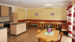 Un comedor con mesas y sillas de madera, una zona de cocina con armarios, y una cortina a rayas rojas y blancas.