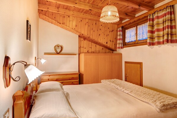 Une chambre avec un plafond en bois, un lit recouvert d’une couette blanche, une commode en bois, une table de chevet, une lampe fixée au mur et une fenêtre avec des rideaux.