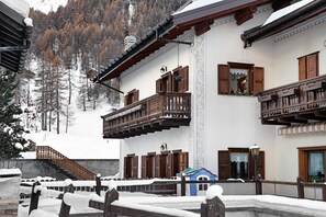Un chalet traditionnel doté de balcons en bois, d’une façade recouverte de neige et surplombant un paysage montagneux.