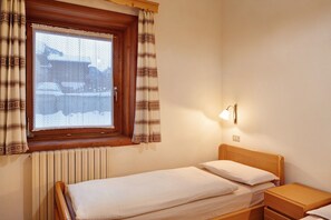 Une petite chambre avec un lit simple, une tête de lit en bois, une table de chevet, une commode, une fenêtre avec des rideaux à motifs et un radiateur.