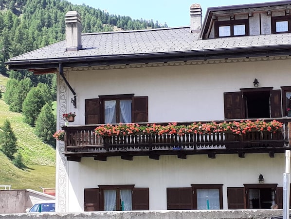 A two-story building with a balcony adorned with flowers, wooden shutters, and a mountainous backdrop.