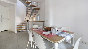 Un comedor con una mesa de madera, sillas blancas, camino de mesa rojo y azul a cuadros, y una escalera con escalones de madera y barandillas de metal.