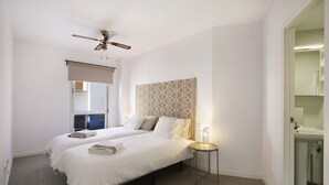 Un dormitorio con dos camas, un ventilador de techo, una mesita de noche con lámpara, y un baño con lavamanos y espejo.