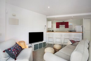 A modern living room with a flat-screen TV, white sofas, and a kitchen area in the background.
