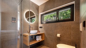 Une salle de bain moderne avec une douche à l’italienne en verre, un lavabo blanc, des toilettes et une fenêtre donnant sur un espace verdoyant.
