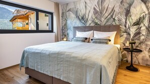 A bedroom with a large bed, a window with a view of a mountain house, and a wall with a leaf pattern. - Apartment "Videre Lodge Wasserfall" with Mountain View, Wi-Fi & Terrace (Gargazon)