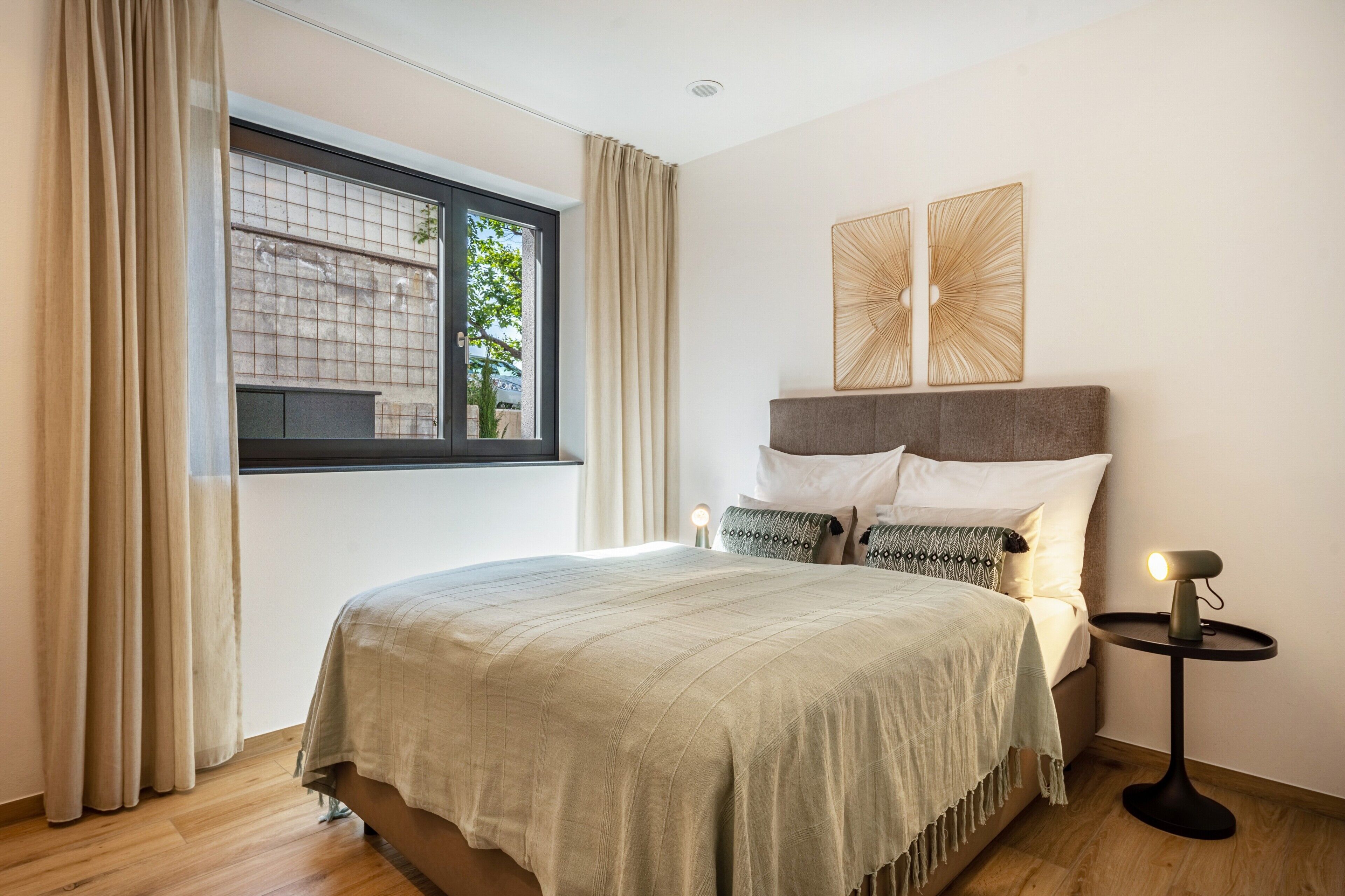 Een slaapkamer met een bed, gordijnen, een raam, een wandlamp en een tafeltje met een lamp.