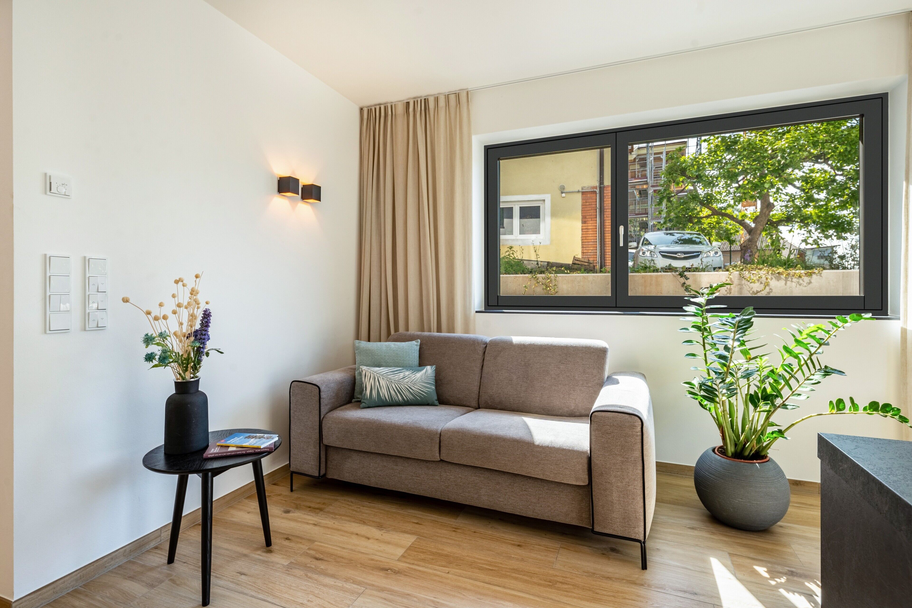 Un salon moderne avec un canapé, une petite table sur laquelle est posé un vase, une plante en pot et une fenêtre donnant sur un arbre et une voiture garée.