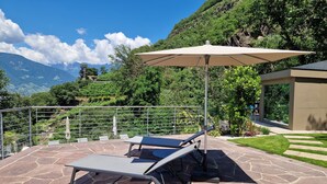 Een terras met twee ligbedden en een grote parasol, een metalen reling en uitzicht op een bergachtig landschap.