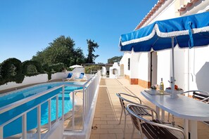 A poolside area with a pool, lounge chairs, a table with a blue umbrella, and a view of the surrounding landscape.