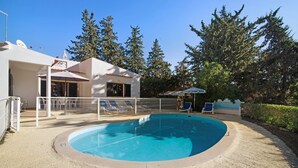 Ein Pool mit einer eingelassenen Liegefläche, Liegestühlen und einem eingezäunten Bereich mit einem Gebäude im Hintergrund.