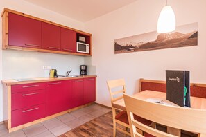 Cocina compacta con gabinetes rojos, microondas y cafetera. Hay una mesa de comedor con sillas y una lámpara colgante. Un cuadro enmarco cuelga de la pared.