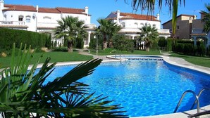 A swimming pool with a clear blue water, surrounded by palm trees and a white building with a red roof in the background.