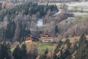 A mountainous landscape with two wooden buildings, one with a sloped roof and another with a flat roof, surrounded by dense forest and a small stream.