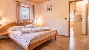 Ein Schlafzimmer mit einem Holzbett, einem fenster mit Vorhängen, einem Nachttisch und Blick in die Küche.