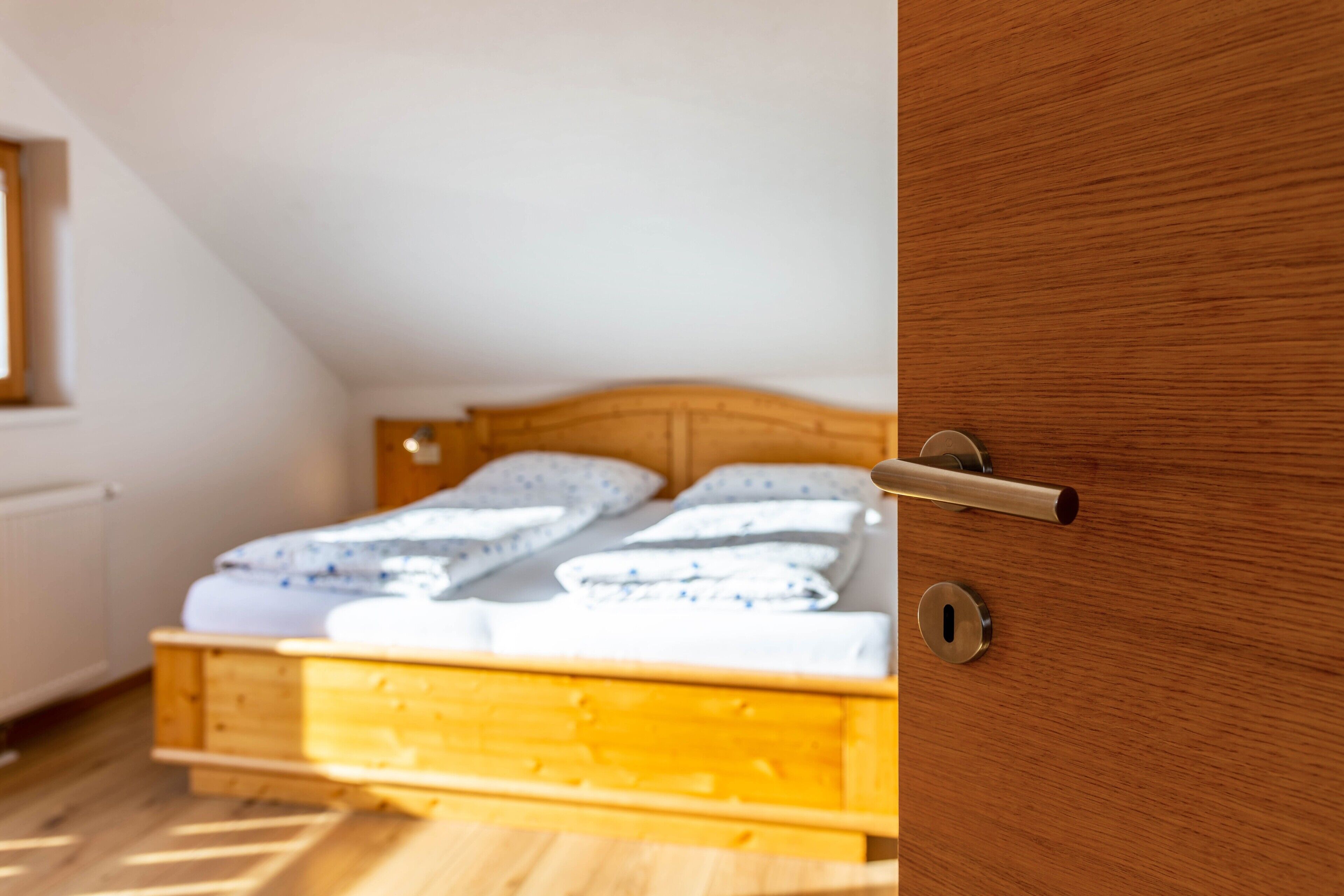 A wooden bed in a room with a sloped ceiling, a door with a handle, and a window with blinds.