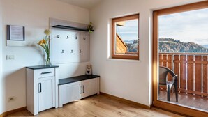 A room with a wooden floor, a white cabinet with drawers, a small table, a chair, a window with a view of hills, and a balcony with a wooden railing. - Apartment 'Kehrerhof - Panorama' with Mountain View, Balcony & Wi-Fi (St. Lorenzen)