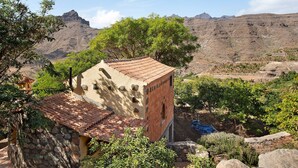A rustic building with a tiled roof, surrounded by greenery and mountains in the background. - Country House "Finca Los Dioses" in the Heart of Nature with Mountain View, Garden & Terrace (Mogán)