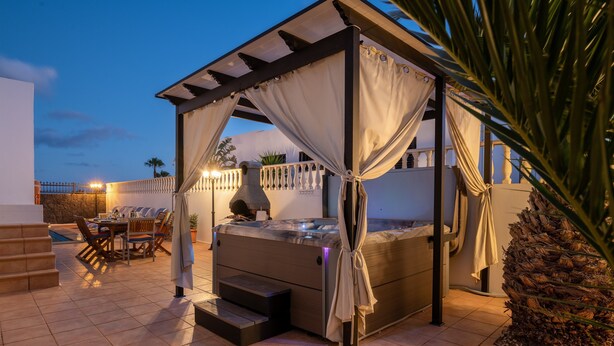 Een overdekt terras met een jacuzzi, een eettafel met stoelen, en een trap naar een zwembad.