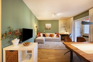 A living room with a flat-screen TV, a sofa with cushions, a dining table, and a window with curtains.