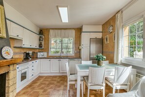 Une cuisine avec des meubles blancs, une table à manger avec des chaises, un four encastré et une fenêtre avec des rideaux.
