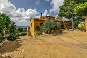 A yellow villa with a tiled roof, surrounded by greenery and a dirt driveway.