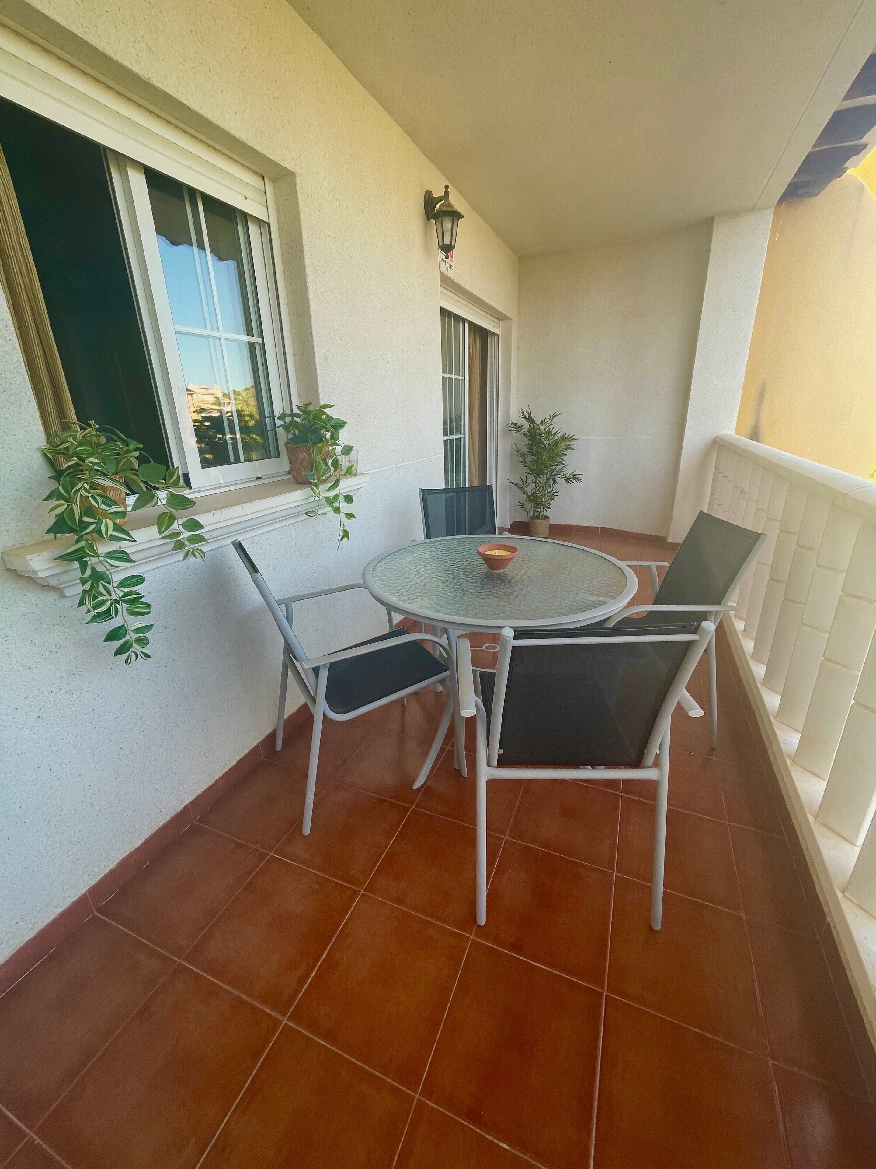 A balcony with a round table and chairs, potted plants, and a window with curtains.