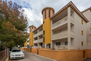 A multi-story building with balconies, a car parked in front, and a tree to the left.