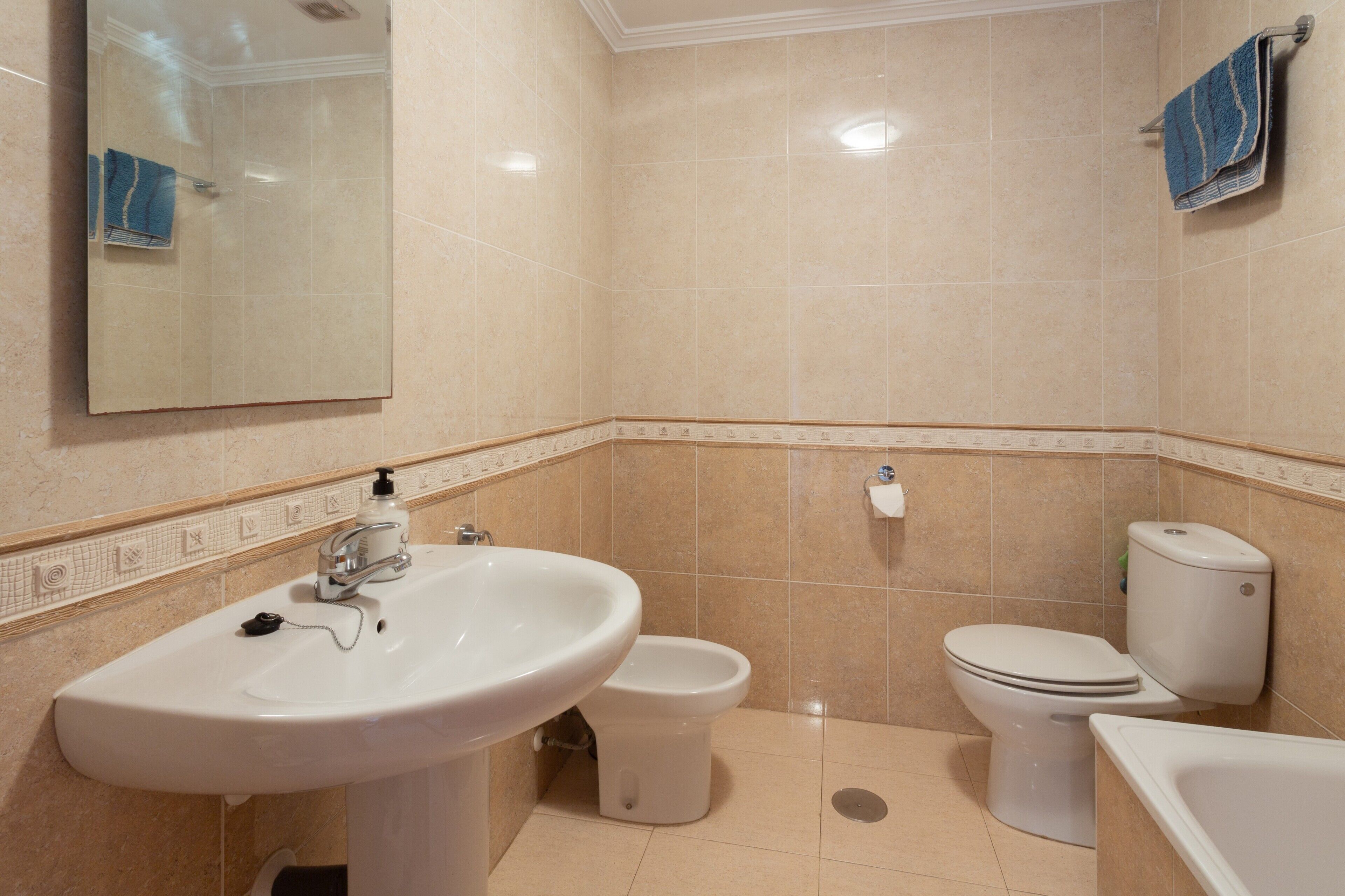 Un bagno con lavandino, WC e vasca da bagno, piastrellato in beige, con uno specchio sopra il lavandino.