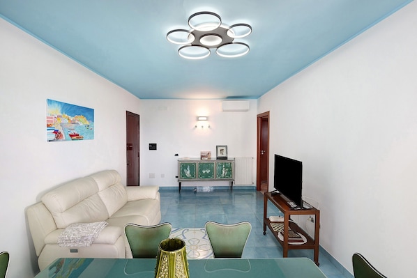 A living room with a sofa, dining table, chairs, a TV on a stand, and a ceiling light fixture.